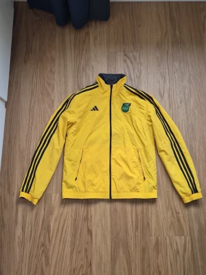 Jamaica Adidas gul reversible träningsjacka x Wales Bonner  - Gul träningsjacka från Adidas med svarta detaljer och tre svarta ränder längs ärmarna. Jackan är reversible och har 2 olika färger har hög krage, två fickor med dragkedja och Jamaica Football Federation-emblem på bröstet. Perfekt för sportaktiviter eller utomhus när det lite blåsigt 