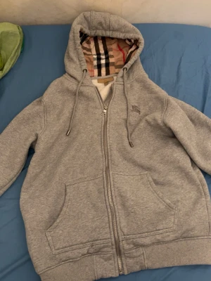 Grå zip hoodie från Burberry - Säljer en grå hoodie från Burberry med dragkedja och klassiskt rutigt foder i huvan. Hoodien har två stora fickor fram, ribbade muddar och justerbar dragsko i huvan. Perfekt för dig som gillar stilrena och exklusiva plagg. Materialet är mjukt och bekvämt.