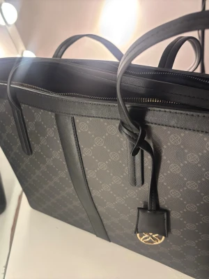 Svart handväska med mönster och logga - Stilren svart handväska från Anna field med diskret mönster och guldfärgad logga. Väskan har två handtag, dragkedja upptill och detaljer i svart läder. Perfekt för dig som gillar minimalistisk design och exklusiva material.