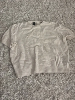 Beige stickad t-shirt från H&M - Säljer en enkel och stilren beige stickad t-shirt från H&M. Tröjan har korta ärmar, rund halsringning och ribbstickad nederkant. Perfekt för dig som gillar en clean och avslappnad look. Materialet är mjukt och lätt.