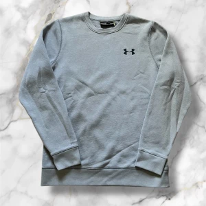 Grå sweatshirt från Under Armour - Säljer en grå sweatshirt från Under Armour med rund halsringning och svart logga på bröstet. Tröjan har långa ärmar och ribbade muddar vid ärmslut och nederkant. Perfekt för träning eller vardag.