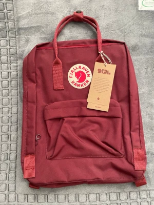 Vinröd Kånken ryggsäck från Fjällräven - Vinröd Kånken ryggsäck från Fjällräven med klassisk rund logotyp framtill. Ryggsäcken har ett stort huvudfack, en mindre frontficka och två handtag upptill. Materialet är slitstark syntet och designen är fyrkantig med justerbara axelband.