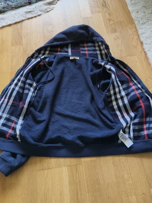 Mörkblå hoodie från Burberry - Snygg mörkblå hoodie från Burberry med dragkedja framtill och klassiskt rutigt foder i huvan. Hoodien har fickor på magen och ribbade muddar vid ärmslut och nederkant. Perfekt för dig som gillar stilrena och exklusiva plagg.