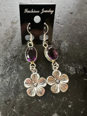 Lila blomörhängen  - Snygga örhängen med stora lila, facetterade stenar och silverfärgade blommor. Hängande modell med krokfäste, perfekt för dig som gillar unika accessoarer med lite extra detaljer. Blommorna har en cool retrostil och stenarna ger ett lyxigt intryck.