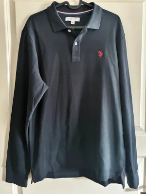 Svart långärmad pikétröja US Polo Assn - Klassisk svart långärmad pikétröja från US Polo Assn med krage och två knappar framtill. Tröjan har en röd broderad logga på bröstet och är tillverkad i mjukt bomullsmaterial. Perfekt för en stilren och avslappnad look.