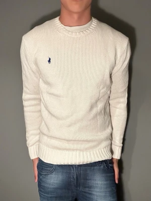 Vit stickad tröja från Polo Ralph Lauren - Säljer en stilren vit stickad tröja från Polo Ralph Lauren med klassisk blå logga broderad på bröstet. Tröjan har rund hals, ribbade muddar och är gjord i mjukt material som passar perfekt till jeans.