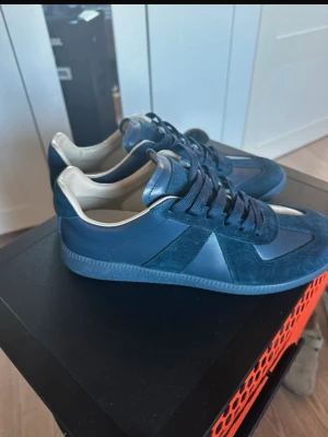 Mörkblå Maison Margiela skor Storlek 43 - Snygga mörkblå sneakers med ovandel i både mocka och slätt läder. Skorna har klassisk låg silhuett, matchande mörkblå snören och beige detaljer vid hälen. Yttersulan är i gummi med mönstrad undersida för bra grepp. Perfekta för en clean och stilren look. Storlek 43, inte använda i nyskick. Pris kan diskuteras, hör av dig 🤙  Ni vet varför priset är som det är . 👍