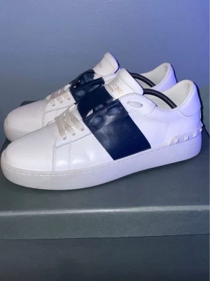 Valentino Garavani vita sneakers - Snygga vita sneakers från Valentino Garavani med en bred mörkblå läderdetalj över snörningen och ikoniska nitar på hälen. Skorna har klassisk låg profil och är tillverkade i premiumläder med vita skosnören och logotyp på tungan.