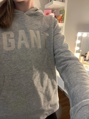 Grå hoodie från GANT - Snygg grå hoodie från GANT med broderad logga och texten 'GANT New Haven' på bröstet. Hoodien har en klassisk huva och en stor magficka framtill. Tillverkad i mjukt sweatshirtmaterial som är perfekt för en avslappnad stil. Inköpspris ca 899