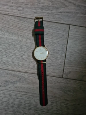 Axcent klocka med randigt band - Stilren klocka från Axcent med ett klassiskt randigt band i grönt och rött. Urtavlan är vit med guldiga detaljer och boett. Bandet är i syntetmaterial och har ett enkelt spänne. Perfekt för dig som vill ha en snygg accessoar till din outfit.