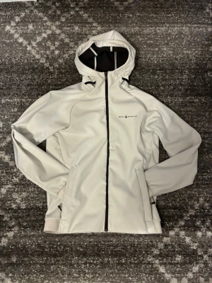 Vit vindjacka från Sail Racing - Snygg vit vindjacka från Sail Racing med svart dragkedja och logga på bröstet. Jackan har huva, två fickor med dragkedja och elastiska muddar vid ärmsluten. Perfekt för blåsiga dagar och sportiga outfits. Tecken på användning.