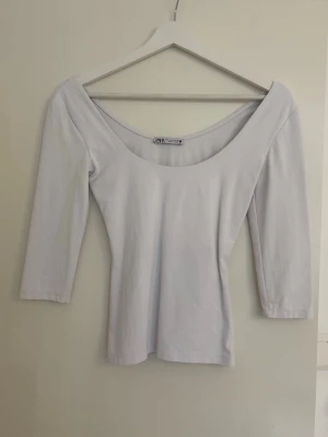 Vit topp med djup urringning från Zara - Vit topp från Zara med djup rund urringning och trekvartsärm. Toppens passform är figurnära och den är tillverkad i ett mjukt bomullsmaterial. Perfekt att styla med jeans eller kjol för en clean look.