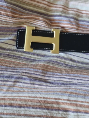 Svart skärp med guldspänne från H - (Helt nytt och oanvänt)Snyggt svart skärp från H med ett stort H-format spänne i guld. Skärpet har vita sömmar längs kanterna som ger en clean kontrast. Perfekt accessoar för att lyfta vilken outfit som helst.