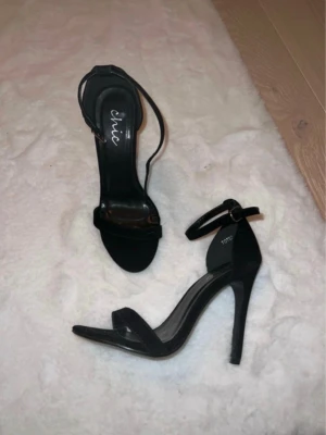 Svarta höga pumps med ankelrem - Säljer ett par svarta pumps från Chic med smal ankelrem och öppen tå. Skorna har hög klack och en stilren design med enkel rem över tårna. Perfekta för dig som gillar klassisk och elegant stil.