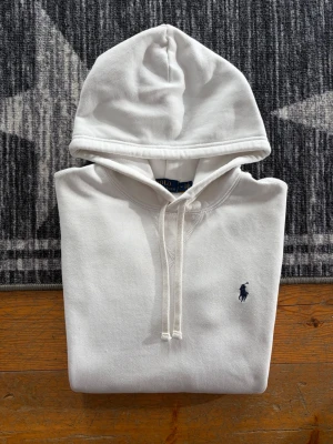 Vit Polo Ralph Lauren Fleece Hoodie- Storlek M - Hej! 👋 Säljer en riktigt fräsch vit Polo Ralph Lauren hoodie i storlek M med den klassiska mörkblå pony-loggan 🐎 Tröjan är 100% äkta med både tvättlappar och QR-kod kvar för enkel verifiering ✅ Den är i mycket fint skick, men jag vill vara ärlig med att det finns två minimala fläckar som knappt syns (se de två sista bilderna för närbild). Det är inget som märks när man bär den! ✨ Ett stilrent basplagg som lyfter vilken outfit som helst. Fraktar snabbt! 🚛📦💬💯 