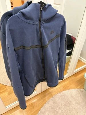 Nike tech fleece mörkblå  - Snygg blå tech fleece från Nike med hel dragkedja och huva. Har en svart vågig detalj över bröstet och diskret Nike-logga på vänster sida. Tillverkad i mjukt sweatshirtmaterial, perfekt för chill eller träning. Byxorna är storlek L och tröjan är M.