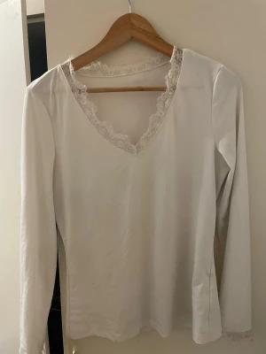 Vit långärmad topp med spets - Vit långärmad topp med v-ringning och fina spetsdetaljer runt halsen och ärmsluten. Enkel och stilren design som passar perfekt till jeans eller kjol. Mjuk och skön, perfekt för dig som gillar en clean look.