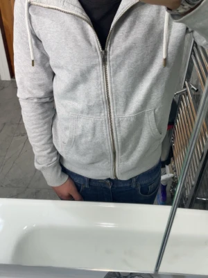 Ljusgrå zip-hoodie med huva - Ljusgrå hoodie med dragkedja och huva, perfekt för chill dagar. Modellen har två stora fickor framtill och snören i huvan. Materialet ser ut att vara mjukt och bekvämt, troligen bomullsblandning. Passar till jeans och sneakers för en avslappnad look.