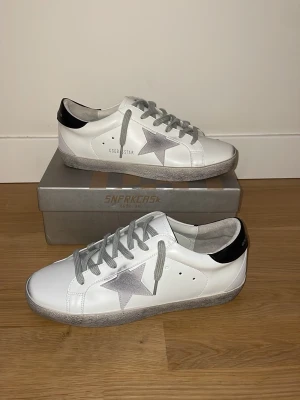 Golden Goose skor - Snygga vita sneakers från Golden Goose med grå snörning och en silverfärgad stjärna på sidan. Skorna har svart detalj på hälen och en slitstark sula i grått. Tillverkade i läder och har en klassisk låg profil som passar perfekt till jeans eller shorts.