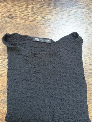 Svart ribbad topp från Zara - En svart topp från Zara med ribbad struktur och korta ärmar. Materialet är syntet och toppen har en enkel, loose passform med rund halsringning. Perfekt för dig som gillar stilrena plagg med lite textur.