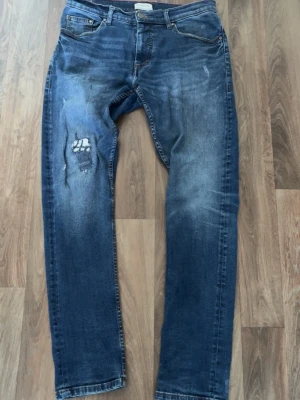 Blå slitna jeans med slitningar - Säljer ett par blå jeans med slitningar och ett tydligt hål på vänster ben. Jeansen har klassisk femficksdesign och raka ben. Materialet är denim och färgen är mörkblå med ljusare tvättade partier. Perfekt för dig som gillar en avslappnad och trendig look.