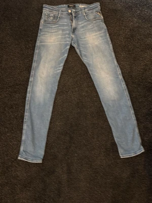 Ljusblå slim jeans från Replay - Säljer ett par ljusblå slim fit jeans från Replay med klassisk femficksdesign och snygga slitningar. Jeansen har normal midja och är tillverkade i bomull med lite stretch för extra komfort. Perfekta för en avslappnad och trendig look.