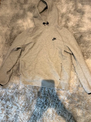 Nike tech hoodie - Snygg grå hoodie från Nike med svart broderad logga på bröstet. Klassisk modell med huva och dragsko, samt fickor på sidorna. Perfekt för en avslappnad och sportig stil.