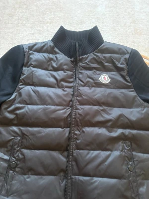 Svart pufferjacka från Moncler - Snygg svart pufferjacka från Moncler med quiltad kropp och släta svarta ärmar. Jackan har ribbad hög krage, dragkedja framtill och två fickor med knappstängning. Ikonisk Moncler-logga på bröstet. Perfekt för kalla vinterdagar.