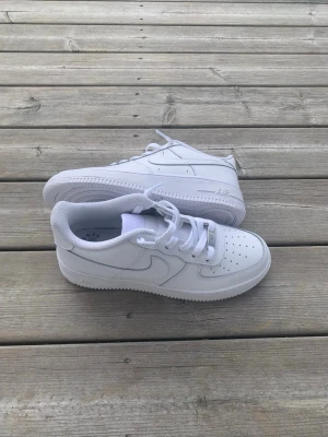 Nike Air Force 1 vita sneakers - Klassiska vita Nike Air Force 1 sneakers i lågt utförande. Skorna har en stilren design med perforerad tå, vit sula och ikoniska Swoosh-loggan på sidan. Tillverkade i läder med textildetaljer och vita skosnören. Perfekta för en clean och tidlös look.