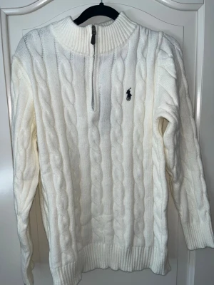 Vit stickad Polo Ralph Lauren tröja - Vit stickad tröja från Polo Ralph Lauren med halv dragkedja och hög krage. Tröjan har det klassiska broderade logotypen på bröstet och ribbade muddar vid ärmslut och nederkant. Perfekt för dig som gillar stilren och tidlös design.