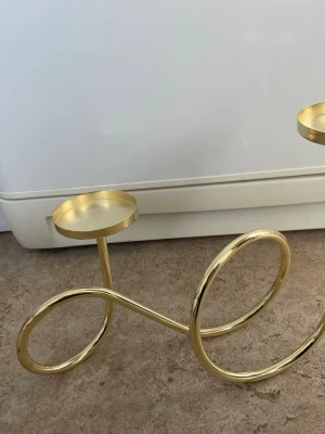 Modern ljusstake i guld metall - Säljer en unik och stilren ljusstake i guld med två hållare för värmeljus. Designen är modern med böjda och slingrande former, tillverkad i metall med blank finish. Perfekt som dekorativ detalj i hemmet.