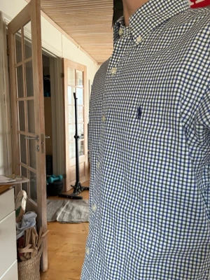Rutig skjorta från Polo Ralph Lauren - Säljer en klassisk långärmad skjorta från Polo Ralph Lauren med blåvitt rutigt mönster och button-down krage. Skjortan har broderad logga på bröstet och knappar framtill. Perfekt för dig som gillar stilrena och tidlösa plagg.