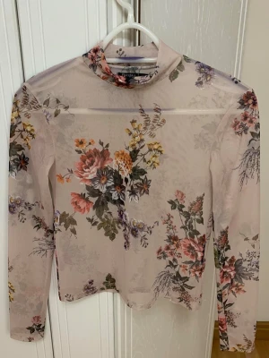 Blommig mesh polotopp från Bershka  - Säljer en transparent, ljusbeige mesh polotopp från Bershka med blommigt mönster i rosa, lila, grön och gul. Toppen har lång ärm och tight passform, perfekt för att styla lager på lager eller som statement-piece.
