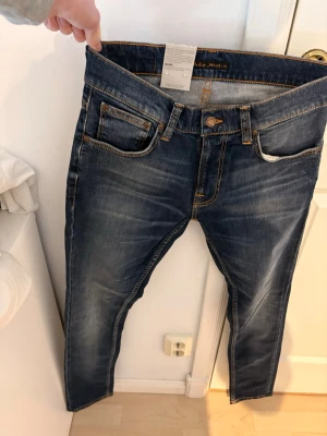 Mörkblå jeans från Nudie Jeans - Säljer ett par mörkblå jeans från Nudie Jeans med snygga slitningar och kontrastsömmar. Klassisk femficksmodell med orange detaljer på bakfickorna och patch med logga bak i midjan. Tillverkade i bomull och har en slim fit passform.