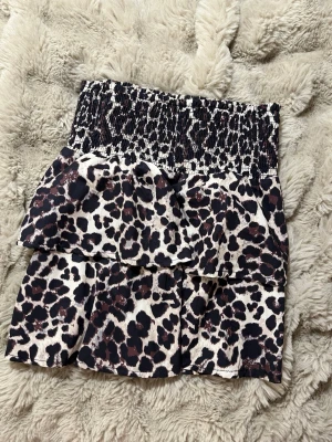 Leopardmönstrad volangkjol - Kort kjol från Instinet med leopardmönster i svart, beige och bruna toner. 100% polyester.  Kjolen har bred smock i midjan och dubbla volanger för en cool och trendig look. Perfekt för dig som vill sticka ut med ett leopardmönster🐆💕!  