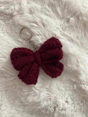 Vinröd virkad rosett nyckelring - Söt handgjord nyckelring med en stor vinröd virkad rosett. Perfekt att hänga på väskan eller nyckelknippan för en personlig touch. Metallring och kedja i silverfärg. Rosetten är mjuk och fluffig, vilket ger en mysig känsla.