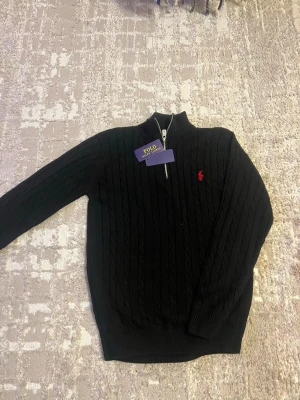 Svart kabelstickad tröja Polo Ralph Lauren - Kabelstickad svart tröja från Polo Ralph Lauren med halv dragkedja och röd broderad logga på bröstet. Tröjan har ribbade muddar och hög krage, perfekt för en stilren och klassisk look.