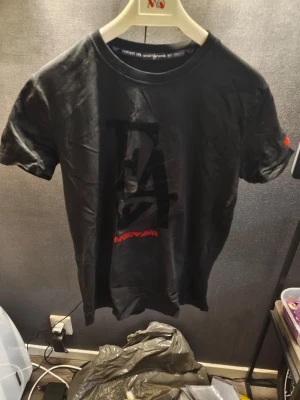 Svart EA t-shirt från Emporio Armani Helg REA NU! - Snygg svart t-shirt från Emporio Armani med stor EA-logga i svart på bröstet och röd textdetalj. Klassisk rund halsringning och korta ärmar. Perfekt för dig som gillar stilrena märkeskläder med en modern touch.
