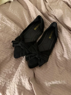 Svarta ballerinaskor med rosett - Stilrena svarta ballerinaskor från Fashion med stor dekorativ rosett framtill. Skorna har fyrkantig tå och är tillverkade i ett mjukt material som liknar mocka. Perfekta för dig som vill ha en klassisk men trendig look.