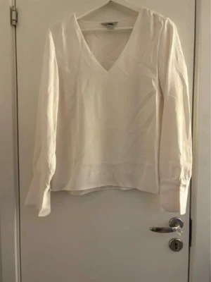 Vit v-ringad blus från H&M - Stilren vit blus från H&M med v-ringning och långa ärmar. Blusen har en avslappnad passform och breda muddar vid ärmsluten. Perfekt att matcha med jeans eller kjol för en enkel och snygg look.