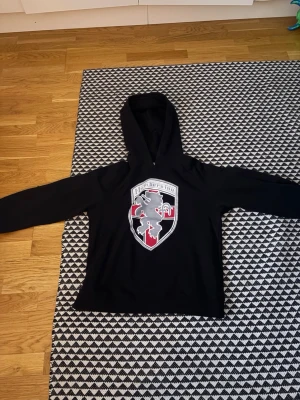 Svart hoodie Numbernine med lejontryck - Svart hoodie från Numbernine med stort tryck av ett lejon och röd korsdetalj på bröstet. Klassisk huva och lång ärm, perfekt för en streetwear-look. Materialet känns mjukt och bekvämt.