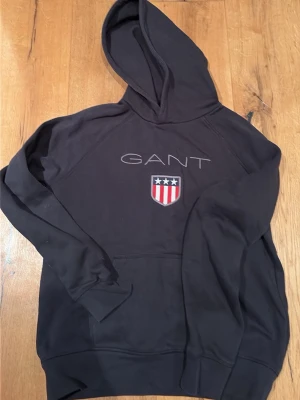 Svart hoodie från GANT med logga - Snygg svart hoodie från GANT med stor huva och klassisk logga broderad på bröstet. På framsidan finns även ett emblem med stjärnor och ränder i rött, vitt och blått. Hoodien har känguruficka och ribbade muddar. Perfekt för en avslappnad och stilren look.