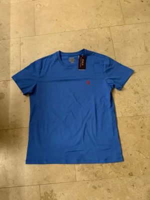 Blå t-shirt från Polo Ralph Lauren - Snygg blå t-shirt från Polo Ralph Lauren med klassisk röd broderad logga på bröstet. T-shirten har rund halsringning och korta ärmar. Tillverkad i mjuk bomull som känns skön mot huden. Perfekt för en clean och stilren look.