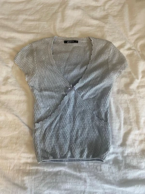 Ljusblå stickad omlottopp från Gina Tricot - Supersöt ljusblå omlottopp från Gina Tricot med korta ärmar och v-ringning. Toppen har en ribbad kant och ett diskret strukturmönster i tyget. Perfekt att styla med jeans eller kjol för en fräsch look. Aldrig använd.