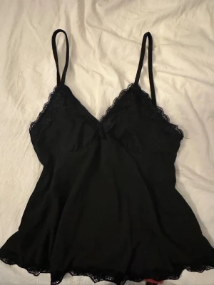 Svart spetsdetaljerad linnetopp - Snygg svart linnetopp med tunna axelband och spetsdetaljer längs urringningen och nederkanten. Mjuk och lätt i materialet, perfekt för att styla lager på lager eller bära som den är. Ingen synlig logga eller märke.