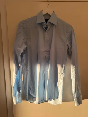 Ljusblå slim fit skjorta Dressmann - Säljer en ljusblå skjorta från Dressmann i slim fit-modell. Skjortan har klassisk krage, långa ärmar och knappar framtill. Perfekt för dig som gillar en stilren och enkel look. Tillverkad i bomull för skön känsla.