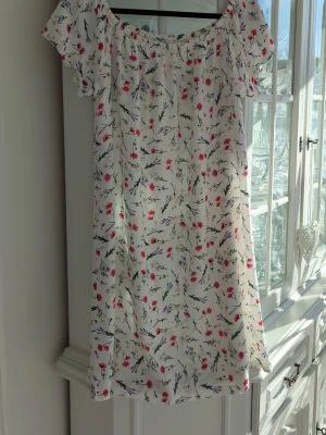 Blommig offshoulder klänning 157 - Söt kort blommig klänning från 157 med offshoulder-modell och korta ärmar. Vit botten med små blommor i rött, lila och grönt. Perfekt för varma dagar och har en luftig, avslappnad passform.