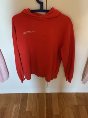 Röd hoodie med tryck framtill - Säljer en röd hoodie med huva och ett litet vitt tryck på bröstet. Hoodien har en avslappnad passform och ribbade muddar vid ärmar och nederkant. Perfekt för en casual och trendig look.