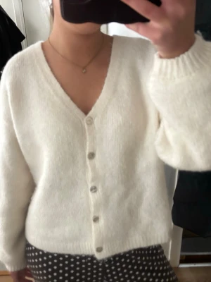 Vit stickad kofta med v-ringning - Mysig vit stickad kofta med v-ringning och knappar framtill. Koftan har långa ärmar och ribbade muddar vid ärmslut och nederkant. Perfekt att slänga över en topp för en chill look.