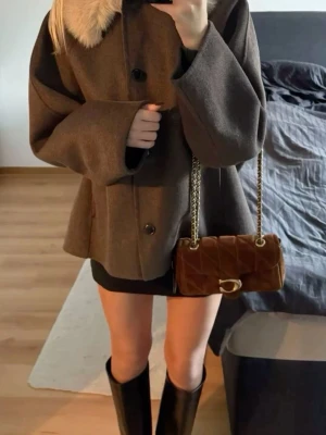 Brun kappa med fuskpäls - Stilren brun kappa med lös passform och stora svarta knappar framtill. Kragen är dekorerad med ljus fuskpäls som ger en lyxig touch. Kappan har även lite mer vida ärmar. Köpt för 1300